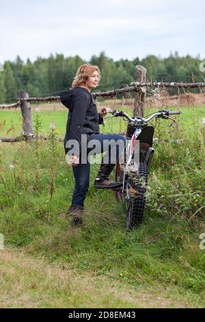 Mature countrywoman standing with dirt motorcycle on rural road near fence Stock Photo