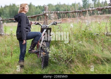 Mature joyful countrywoman standing with dirt motorcycle on rural road, copy space Stock Photo