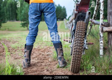 Male rider well shod in protective boots standing near his off-road motorcycle on dirt road, rear view Stock Photo