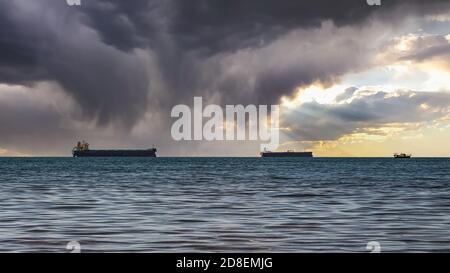 Close-up of a cloudy seascape and a bright sunset. Blue sea, storm clouds with sun rays over the horizon, and several cargo ships Stock Photo