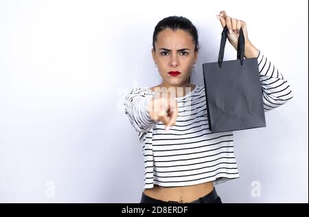 Young beautiful woman holding a black shopping bag pointing with finger to the camera and to you, hand sign, gesture from the front. Black Friday Stock Photo
