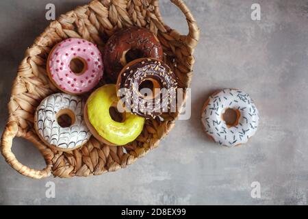 Sweet and fresh colored different donuts with chocolate frosted, glazed and sprinkles, icing topping in wicker basket plate on concrete background. Stock Photo