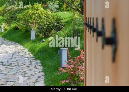 Perspective view through blurred open wooden gate with metal decor to stone walkway in Japanese Zen garden lined with ornamental plants and lanterns  Stock Photo