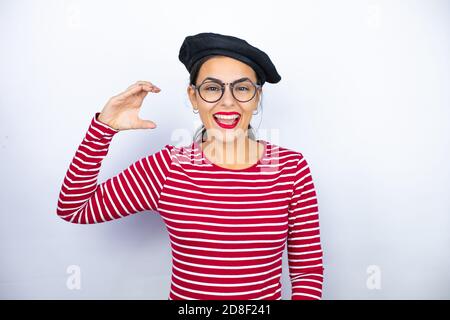 Young beautiful brunette woman wearing french beret and glasses over white background smiling and confident gesturing with hand doing small size sign Stock Photo