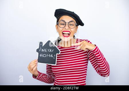 Young beautiful brunette woman wearing french beret and glasses over white background smiling and holding house blackboard with new home word message Stock Photo