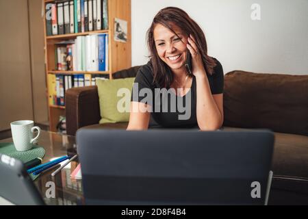 Smiling businesswoman talking on smartphone while using laptop in living room at home Stock Photo