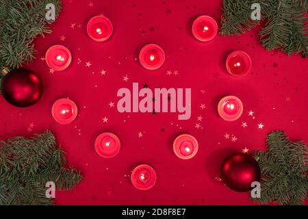 Christmas romantic evening composition made from burning candles, tree branches, glitter and baubles on red surface. Merry christmas and happy 2021 Stock Photo