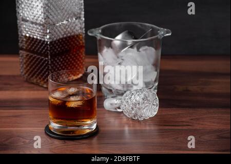 Selective focus on a glass with whiskey and three ice cubes. Defocused ice bucket, cap and ice bucket. Wooden table Stock Photo