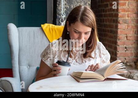 A beautiful young woman with red long hair in a white blouse sits in a chair in cafe , drinks coffee and reads a book Stock Photo