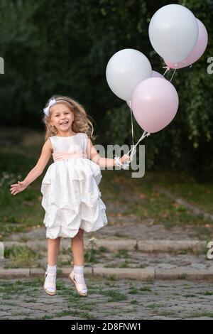 Little girl in white dress with balloons jumping on the street Stock Photo