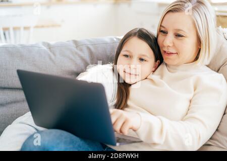 Caucasian mommy with daughter lying on the couch talking and looking at the laptop screen. Stock Photo