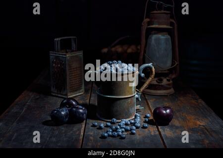 Classic still life composition - blueberry in old copper pot and plums on shabby old wooden table, blurred objects in the dark background - photograph Stock Photo