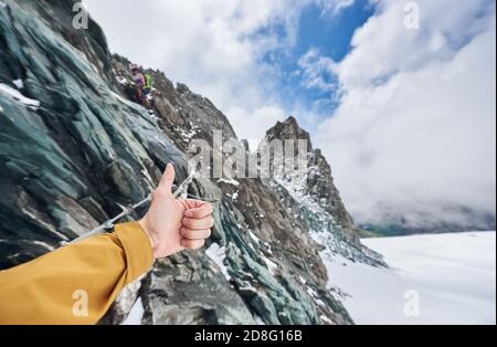 Close up of alpinist hand doing approval gesture with rocky mountains and sky on background, showing thumbs up sign while friend climbing winter mount Stock Photo