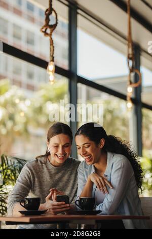 Two women sitting in a restaurant looking at mobile phone. Friends sitting at a cafe with coffee on the table looking at mobile phone. Stock Photo