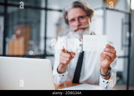 Holding paper with text you are fired. Mature businessman with grey hair and beard in formal clothes is in the office Stock Photo