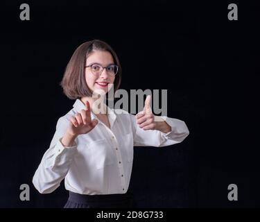 Caucasian girl in a white shirt and glasses shows something in front with her hand, holds her second hand with thumb up Stock Photo