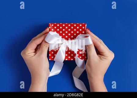 Hands tie a ribbon bow on a red gift box top view on blue background Stock Photo