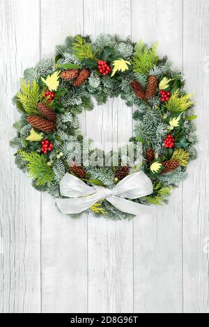 Winter solstice & Christmas  wreath with snow covered fir, cedar, holly, ivy, mistletoe, cedar, pine cones & white bow on rustic wood background. Comp Stock Photo