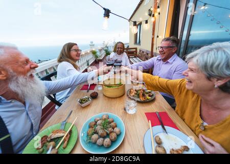 Happy multiracial seniors toasting with red wine glasses together on house patio dinner - Elderly lifestyle people concept