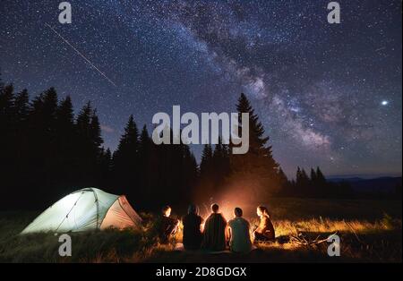 Evening summer camping, spruce forest on background, sky with falling stars and milky way. Group of five friends sitting together around campfire in mountains, enjoying fresh air near illuminated tent Stock Photo