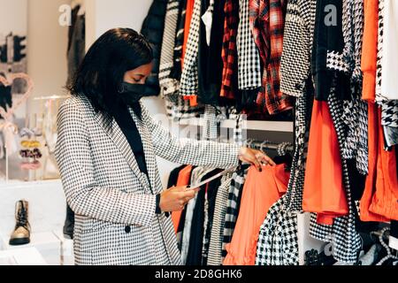 black woman manager in a fashion retail store is wearing a mask as protection against virus and using a tablet for working and checking the shop stock Stock Photo