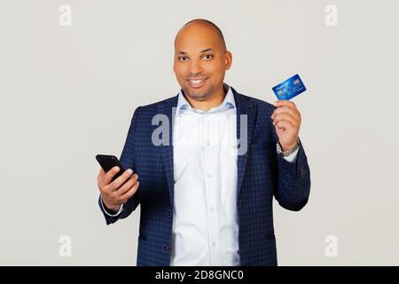 Portrait of happy young African American guy businessman using credit card to pay online using smartphone. Standing on a gray background Stock Photo