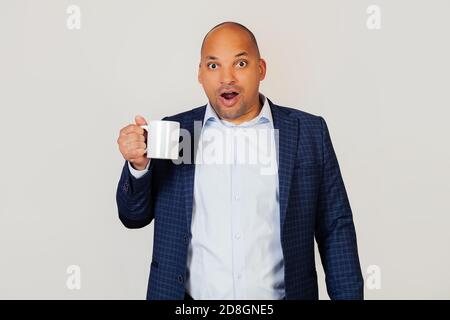 Portrait of astonished young african american businessman guy, drinking a cup of coffee, scared shocked with surprised face, scared and excited with fear expression. Standing on a gray background Stock Photo