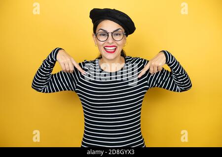 Young beautiful brunette woman wearing french beret and glasses over yellow background looking confident with smile on face, pointing oneself with fin Stock Photo