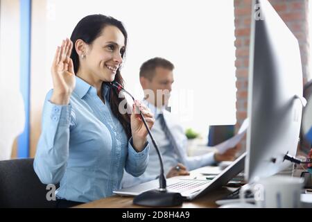 Smiling businesswoman sits at computer screen with microphone and waves hand with colleague in background in office Stock Photo