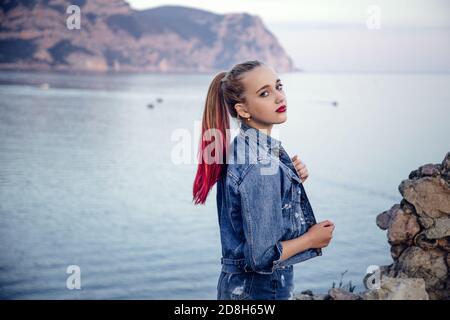 A young girl with bright eyes and lips. She is blonde with partially dyed in pink and stands against the blue sea. Background blurred for artistic pur Stock Photo