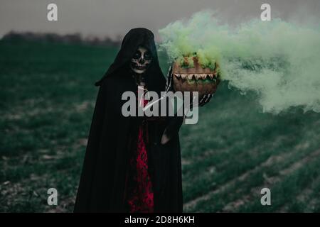 Young woman stands among field in Halloween costume of death with painted skeleton on her body and holds jack-o-lantern in her hands with yellow smoke