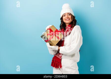 young joyful woman in winter outfit holding wrapped present on blue Stock Photo