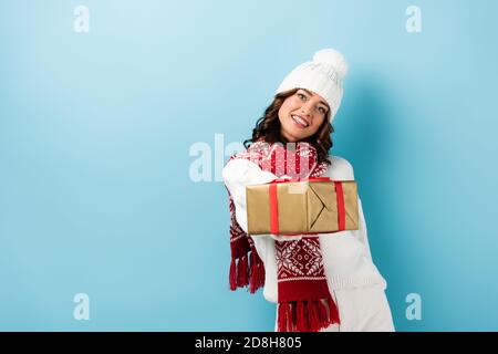 young pleased woman in winter outfit holding wrapped present on blue Stock Photo