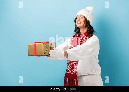 young pleased woman in winter outfit with outstretched hands holding wrapped present on blue Stock Photo