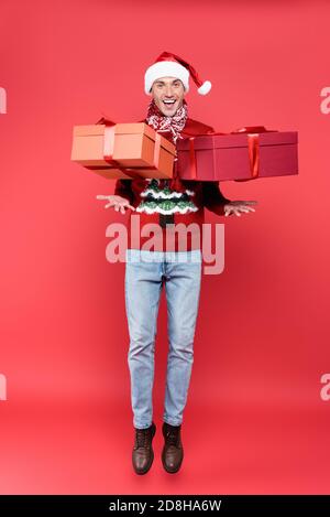 Positive man in santa hat jumping near presents on red background Stock Photo