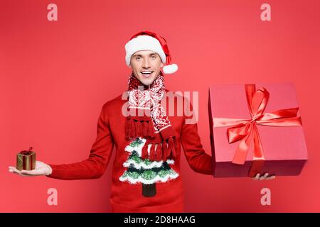 Positive man in sweater and santa hat holding gift boxes on red background Stock Photo