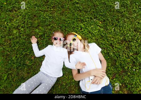 Two small smiling girls lying on the grass in the Park in white t-shirts and headphones Stock Photo