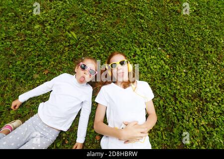Two small smiling girls lying on the grass in the Park in white t-shirts and headphones Stock Photo