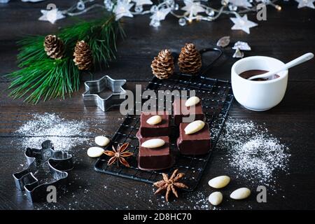 Marzipancubes with chocolate icing on a black cooling grid, decorated with pine branch, cinnamon sticks and aniseed stars, fir cones, next to it a bow Stock Photo