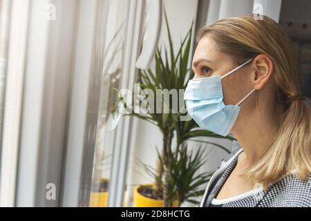 home quarantine - young depressed woman with medical face mask looking through window Stock Photo