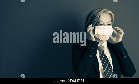 Pensive business woman straightens her face mask standing on gray background with copy space at left. Toned black and white image Stock Photo