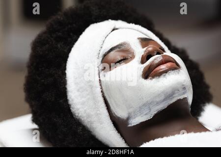 Close up view of curly african american woman with white headband and face mask lying in spa salon Stock Photo