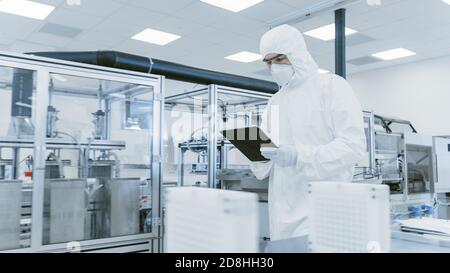 Quality Control Check: Scientist Using Digital Tablet Computer and wearing Protective Suit walks through Manufacturing Laboratory. Product Stock Photo