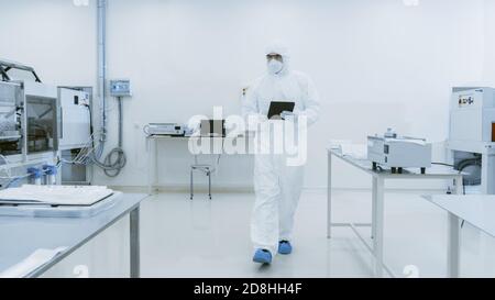 Quality Control Check: Scientist Using Digital Tablet Computer and wearing Protective Suit walks through Manufacturing Laboratory. Product Stock Photo