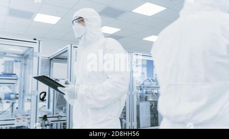 Quality Control Check: Scientist Using Digital Tablet Computer and wearing Protective Suit walks through Manufacturing Laboratory. Product Stock Photo