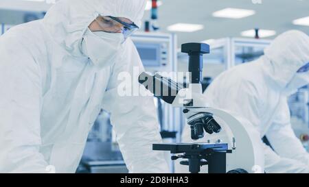 In Laboratory Scientist in Protective Clothes Doing Research, Using Microscope and Writing Down Data. Workers Working on a Modern Manufactory Stock Photo