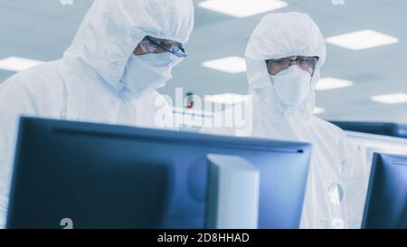 Two Scientists Technicians in Sterile Suits Check Data on Personal Computer, the Have Discussion. They Work in Pharmateutics Manufacturing Laboratory. Stock Photo