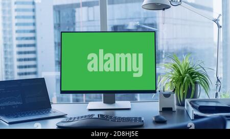 Shot of the Office Desk with Green Mock-up Screen Personal Computer Standing on it. Modern Stylish Room with a Big City Business District View. Stock Photo