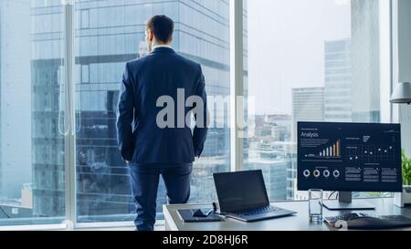 Confident Businessman in a Suit Contemplating Business Deal in His Office, Looking out of the Window. Window Has Panoramic View on Big City Business Stock Photo