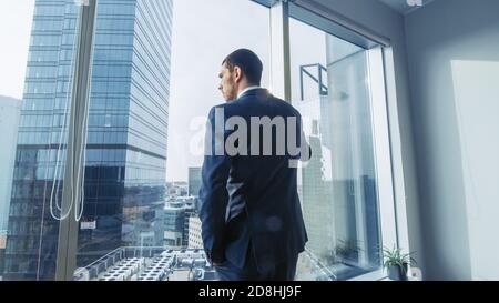 Thoughtful Businessman Wearing Suit Standing in His Office, Looking out of the Window and Contemplating Next Big Business Contract. Major City Stock Photo